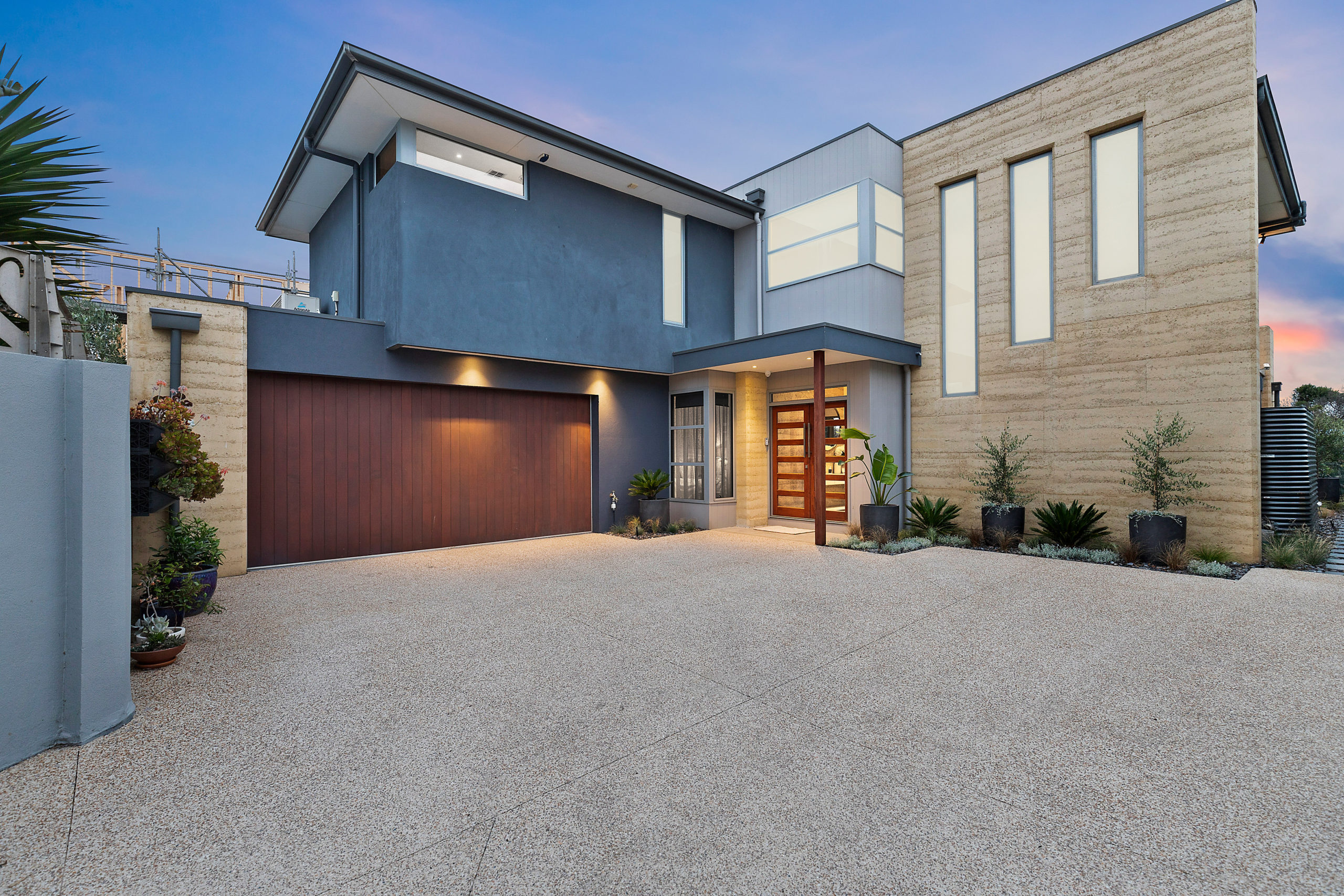 Looking at the front of the home from the driveway, Rammed earth walls are featured. These are complemented by rendered walls and vertical grooved timber panelling. The garage door is in vertical timber look panels to finish of the façade.